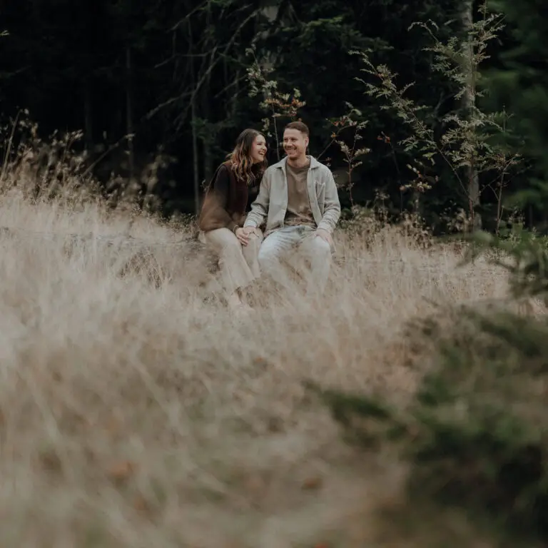 Paarfotos Steiermark von Lisa und Mathieu am Masenberg, beide sitzen nebeneinander auf einem umgestürzten Baumstamm und lachen ungezwungen Feature Image